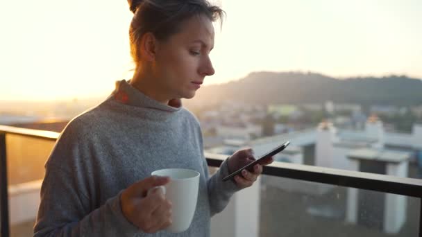 La mujer comienza el día con una taza de té o café y revisando correos electrónicos en su teléfono inteligente en el balcón al amanecer, en cámara lenta. — Vídeo de stock