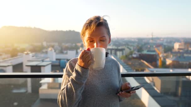 Mulher começa seu dia com uma xícara de chá ou café e verificar e-mails em seu smartphone na varanda ao amanhecer, câmera lenta. — Vídeo de Stock