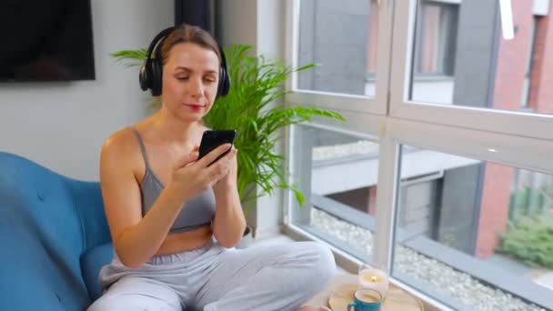 音楽を聴いたり、モバイルアプリを使用したり、自宅のソファに座っているスマートフォン上のソーシャルネットワーク上で通信する無線ヘッドフォンの女性 — ストック動画
