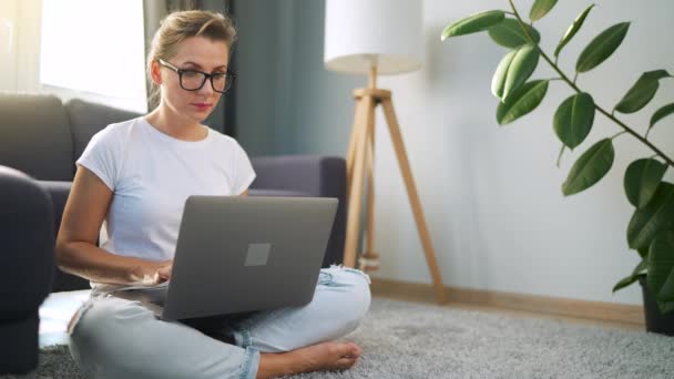 Жінка в окулярах сидить на підлозі і працює на ноутбуці. Концепція віддаленої роботи . — стокове відео