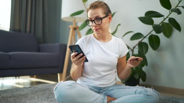 Frau mit Brille sitzt auf dem Boden und kauft online mit Kreditkarte und Smartphone ein. Online-Shopping, Lifestyletechnologie — Stockvideo