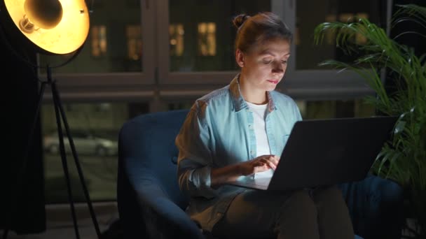女性はアームチェアに座って、夜にラップトップで作業しています。リモートワークの概念. — ストック動画