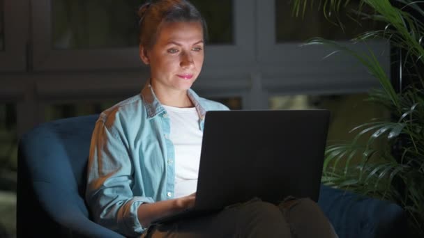 Donna è seduta sulla poltrona e lavora su un computer portatile di notte. Concetto di lavoro a distanza. — Video Stock