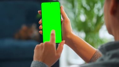 Evde dikey modda yeşil ekranlı akıllı telefon kullanan bir kadın var. Kız internette geziniyor, içeriği izliyor, videolar