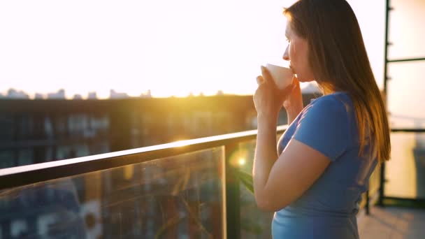 Mulher com uma xícara de café em pé na varanda e admirar o pôr do sol. Movimento lento — Vídeo de Stock