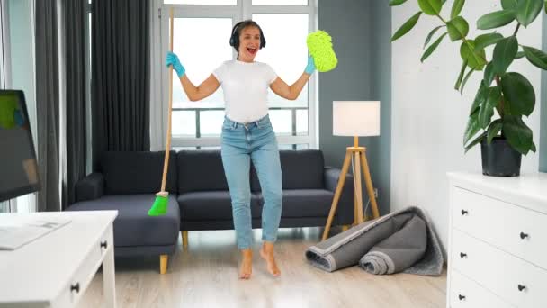 Femme écoute de la musique avec des écouteurs et nettoyer la maison et s'amuser à danser avec un balai et une éponge. Mouvement lent — Video
