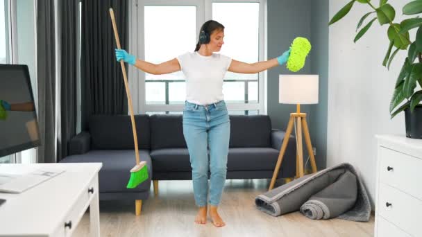 Vrouw luistert naar muziek met koptelefoon en schoonmaken van het huis en heeft plezier met dansen met een bezem en spons. Langzame beweging — Stockvideo