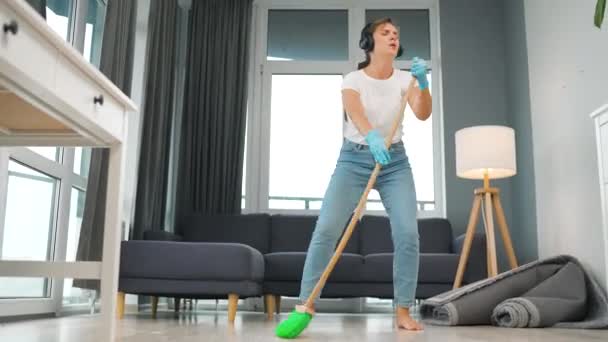 Mujer con auriculares limpia la casa y se divierte cantando con una escoba como una estrella en un concierto. — Vídeo de stock