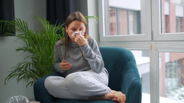 Ongezonde vrouw zit in een stoel en niest of blaast haar neus in een servet omdat ze een verkoudheid, griep, coronavirus heeft. Het sneeuwt buiten. — Stockvideo