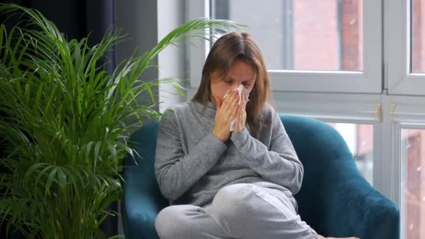 Nezdravá žena sedí v křesle a kýchá nebo se vysmrká do ubrousku, protože má nachlazení, chřipku, koronavirus. Venku sněží. — Stock video