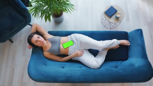Εναέρια λήψη χαλαρής γυναίκας που ακούει διαλέξεις ή μουσική σε ακουστικά σε smartphone με πράσινη οθόνη mock-up, ξαπλωμένη στον καναπέ στο σπίτι. — Αρχείο Βίντεο