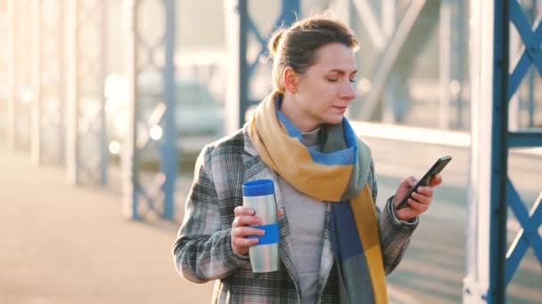 Портрет молодої кавказької бізнес-леді в пальто, що йде через міст в морозний ранок, п'є каву і використовує смартфон. Комунікація, робочий день, концепція зайнятого життя. Повільний рух — стокове відео