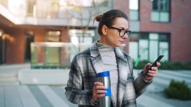Portrait of a young caucasian businesswoman with glasses and a coat walks through the business district, with thermo cup and using smartphone. Communication, work day, busy life concept. — Stock Video
