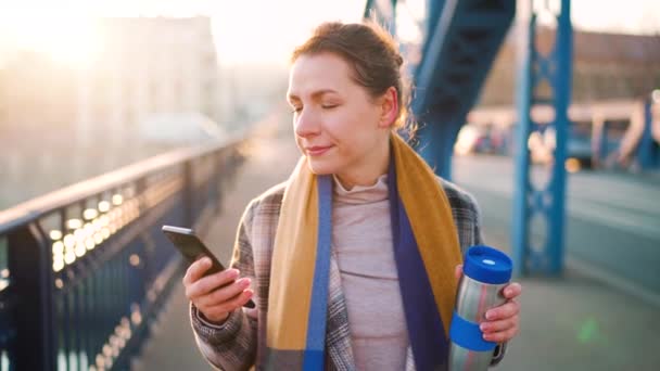 Retrato de una joven mujer de negocios caucásica en un abrigo, caminando por el puente en una mañana helada, bebiendo café y usando un teléfono inteligente. Comunicación, día de trabajo, concepto de vida ocupada. Movimiento lento — Vídeo de stock