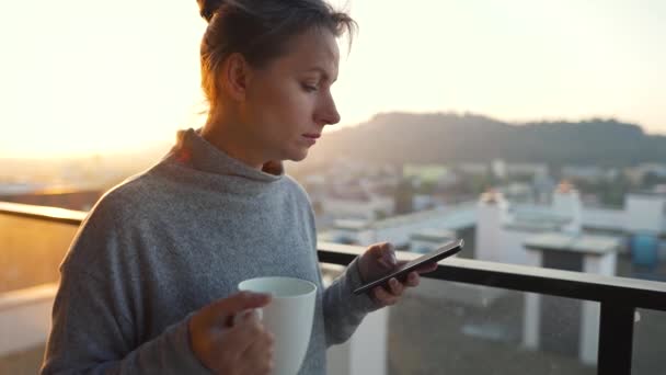 Mulher começa seu dia com uma xícara de chá ou café e verificar e-mails em seu smartphone na varanda ao amanhecer, câmera lenta. — Vídeo de Stock