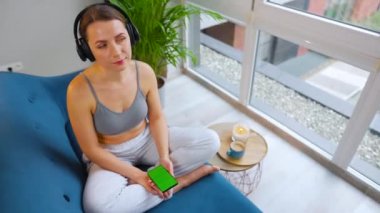 Kablosuz kulaklık takan, akıllı telefondan müzik dinleyen yeşil ekran televizyonlu bir kadın evdeki kanepede oturuyor.