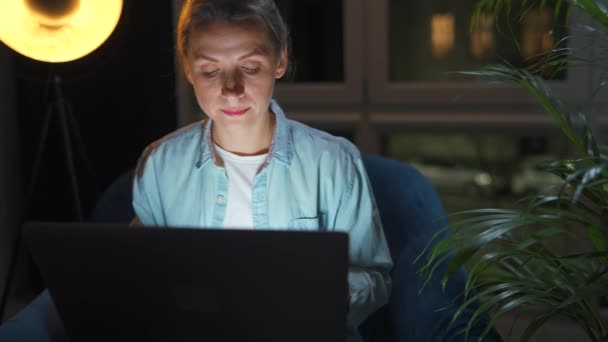 La mujer está sentada en el sillón y trabajando en un portátil por la noche. Concepto de trabajo remoto. — Vídeo de stock