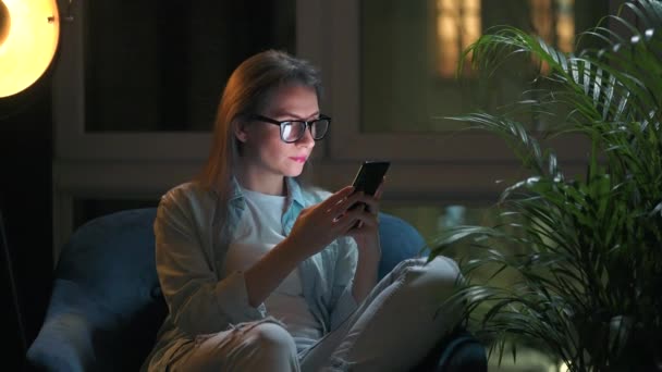 Mujer con gafas está sentado en el sillón y el uso de teléfono inteligente por la noche por la noche. Concepto de trabajo remoto. — Vídeo de stock