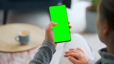 Evde dikey modda yeşil ekranlı akıllı telefon kullanan bir kadın var. Kız internette geziniyor, içeriği izliyor, videolar