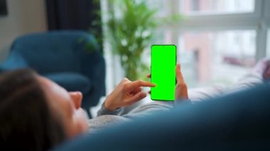 Kadın evde kanepede yatıyor ve dikey modda yeşil maket ekranlı akıllı telefon kullanıyor. Kız internette geziniyor, içeriği, videoları, blogları izliyor.