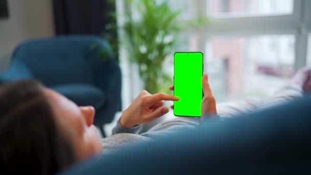 Woman lies at home on the sofa and using smartphone with green mock-up screen in vertical mode. Girl browsing Internet, watching content, videos, blogs. — Stockvideo