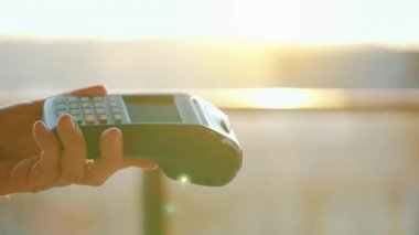 Yakın plan, kadın banka terminalindeki siparişi ödemek için akıllı telefon nakitsiz cüzdan teknolojisi kullanıyor. Kart makinesi erkek eliyle güneş ışığı arkaplanında