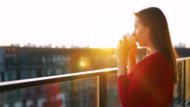 Kırmızı elbiseli bir kadın elinde bir fincan kahveyle balkonda duruyor ve gün batımını seyrediyor. Yavaş çekim — Stok video
