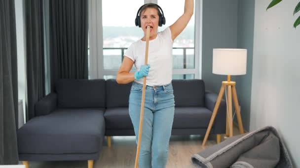 Een vrouw met een koptelefoon maakt het huis schoon en zingt met een bezem als een ster tijdens een concert. Langzame beweging — Stockvideo