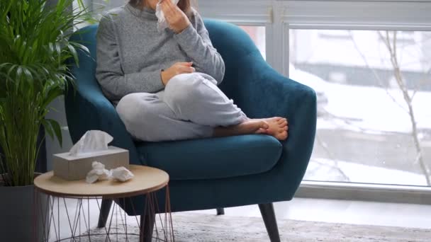 Unhealthy woman sits in a chair and sneezes or blows her nose into a napkin because she has a cold, flu, coronavirus. It is snowing outside — Stock Video