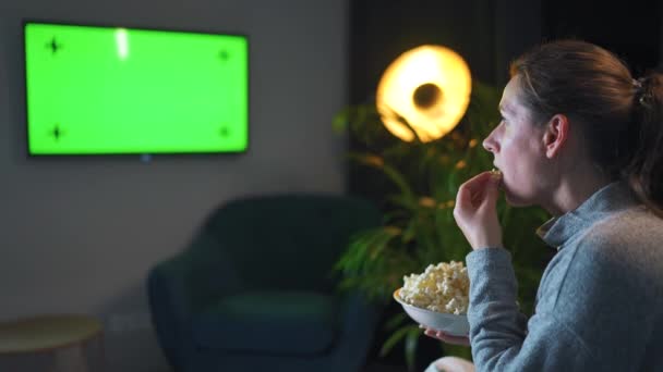Donna seduta su un divano in soggiorno la sera e guardando un modello di schermo verde TV. La donna è infatuata di guardare e mangiare popcorn. — Video Stock