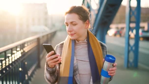 Portrait of a young caucasian businesswoman in a coat, walking across the bridge on a frosty morning, drinking coffee and using smartphone. Communication, work day, busy life concept. — Video Stock