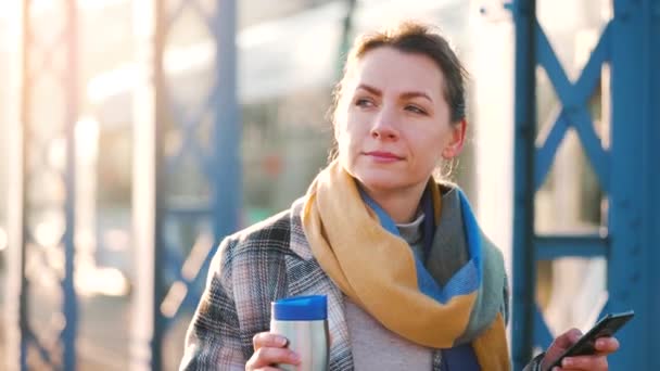 Portret van een jonge blanke zakenvrouw in een jas, rondlopend in de stad op een ijzige ochtend, koffie drinkend en met een smartphone. Communicatie, werkdag, druk levensconcept. Langzame beweging — Stockvideo