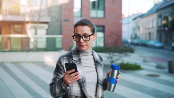 Porträtt av en ung kaukasisk affärskvinna med glasögon och en rock går genom affärsdistriktet, med termo cup och använda smartphone. Kommunikation, arbetsdag, upptagen livskoncept. Långsamma rörelser — Stockvideo