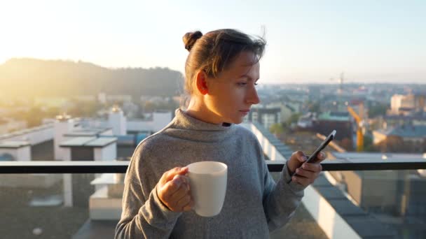 Femme commence sa journée avec une tasse de thé ou de café et vérifier les e-mails dans son smartphone sur le balcon à l'aube, au ralenti. — Video