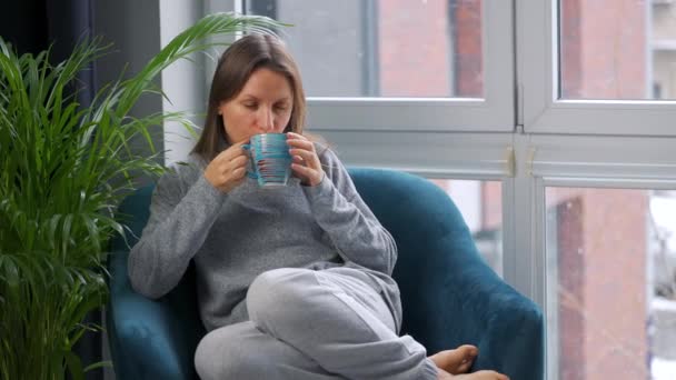 Mujer caucásica poco saludable se sienta en una silla y bebe té caliente o un medicamento para el resfriado porque tiene un resfriado, gripe, coronavirus. Está nevando afuera. — Vídeos de Stock