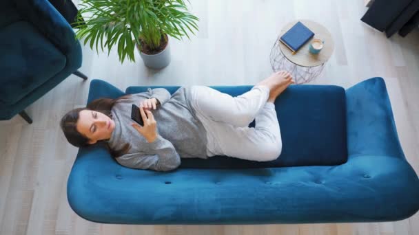 Overhead shot van de vrouw gaat naar de bank en gaat liggen op het, het houden van een smartphone, met behulp van mobiele apps, kijken naar video, chatten in sociale media. — Stockvideo