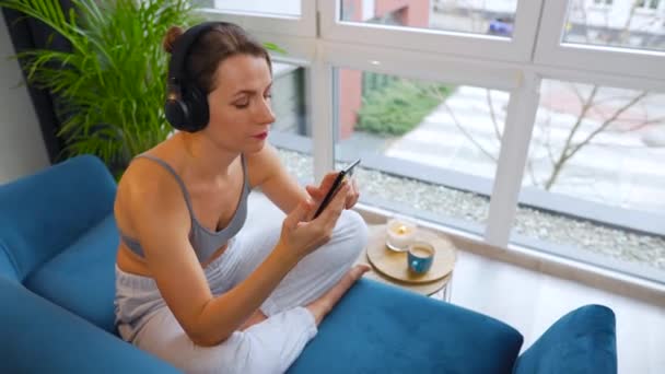 Mulher em fones de ouvido sem fio ouvindo música e usando aplicativos móveis ou se comunica em redes sociais no smartphone sentado no sofá em casa — Vídeo de Stock