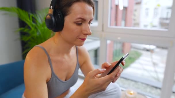 音楽を聴いたり、モバイルアプリを使用したり、自宅のソファに座っているスマートフォン上のソーシャルネットワーク上で通信する無線ヘッドフォンの女性 — ストック動画