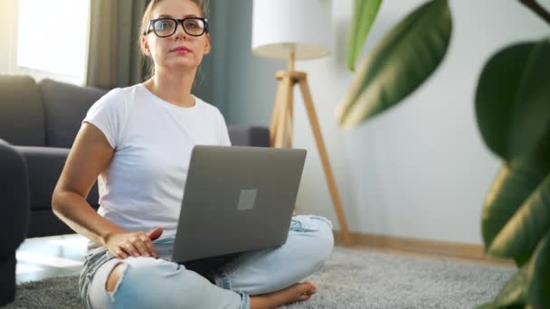Portret van een vrouw met een bril die over een laptop naar de camera kijkt in het interieur van een gezellig appartement. Begrip werk op afstand. — Stockvideo