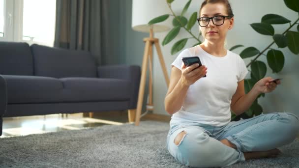 Mulher com óculos está sentado no chão e faz uma compra on-line usando um cartão de crédito e smartphone. Compras online, tecnologia de estilo de vida — Vídeo de Stock