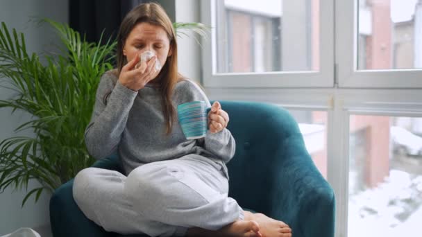 건강하지 못한 여자는 의자에 앉아 뜨거운 차나찬 약을 마시고 감기, 독감, 코로나 바이러스가 있기 때문에 재채기를 하거나 냅킨에 코를 불어넣는다. 밖에 눈 이 내리네 — 비디오
