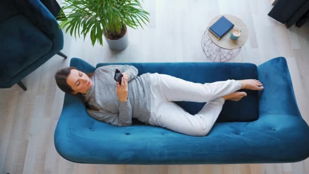 Nahoře záběr šťastné uvolněné ženy držící chytrý telefon, pomocí mobilních aplikací, sledování legračního videa, baví chatování na sociálních sítích, ležící na gauči doma. — Stock video