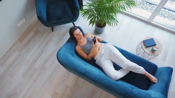 Donna con cuffie wireless che ascolta musica e utilizza app mobili o comunica sui social network su smartphone sdraiata sul divano di casa, vista dall'alto — Video Stock