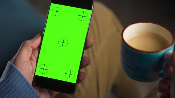 Man använder smartphone med grön mock-up skärm i vertikalt läge och dricka kaffe. Man surfar på Internet, tittar på innehåll, videor. — Stockvideo