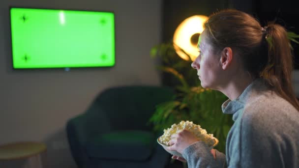 한 여자가 저녁에 거실에 있는 소파에 앉아 녹색 TV 화면을 보고 있습니다. 여자는 팝콘을 보고 먹는 일에 푹 빠져 있다. — 비디오