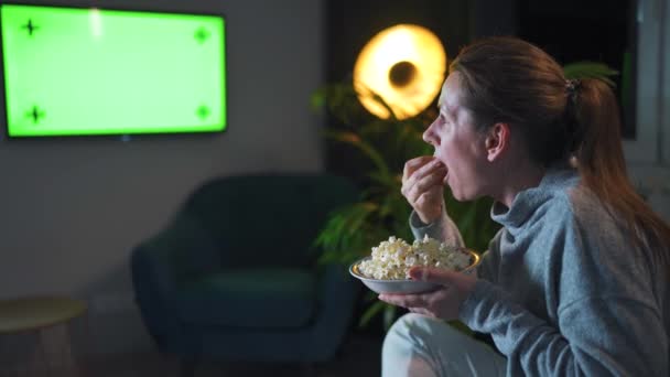 Frau, die abends auf einem Sofa im Wohnzimmer sitzt und eine grüne Fernsehbildschirm-Attrappe anschaut, ist emotional besorgt über das, was sie sieht. Die Frau ist vernarrt darin, Popcorn zu sehen und zu essen. — Stockvideo