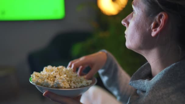 Donna seduta su un divano in soggiorno la sera e guardando un modello di schermo verde TV. La donna è infatuata di guardare e mangiare popcorn. — Video Stock