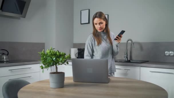 Жінка зі смартфоном в руці, стоячи вдома затишна кухня, використовує ноутбук робити позаштатну роботу, виглядаючи зосередженою глибоко втраченою в думках. Концепція віддаленої роботи. Домашній офіс — стокове відео