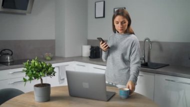 Akıllı telefonlu bir kadın evde rahat bir mutfakta dizüstü bilgisayar kullanıyor, bağımsız çalışıyor, derin düşüncelere dalmış görünüyor. Uzaktan kumanda kavramı. İçişleri Bakanlığı