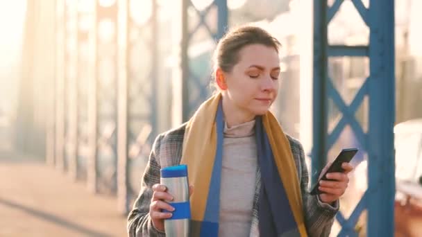 Portrait of a young caucasian businesswoman in a coat, walking around the city on a frosty morning, drinking coffee and using smartphone. Communication, work day, busy life concept. — Stock Video